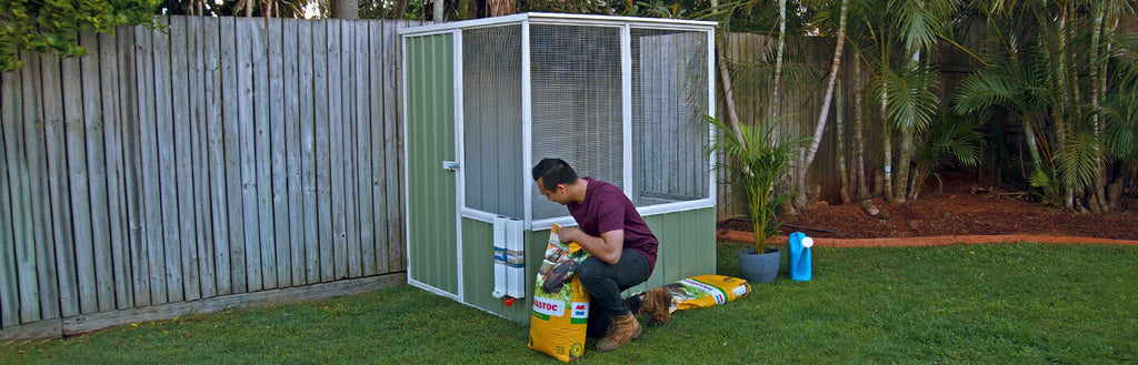 A crouched person replacing the feed on a green, steel chicken coop.